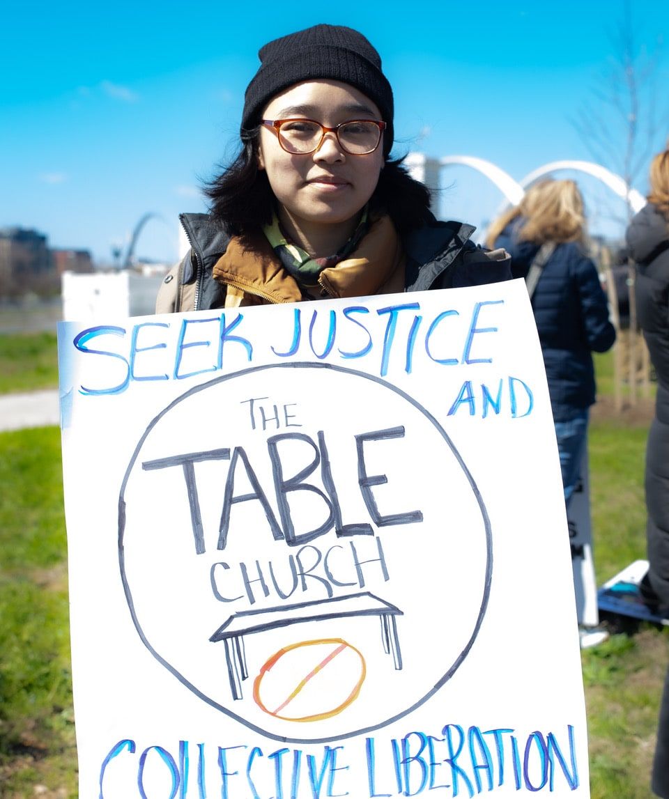 A person holds a hand-lettered Table Church sign reading 'Seek justice and collective liberation' at a march.